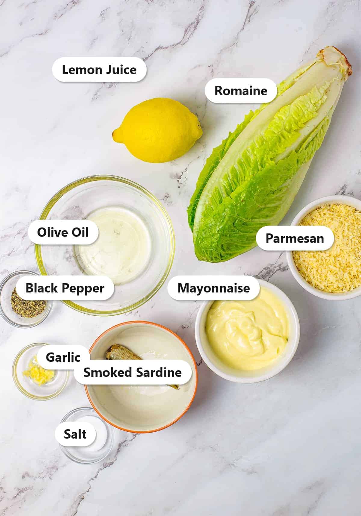 Labeled ingredients for homemade Caesar salad dressing including mayonnaise, smoked sardine, Parmesan cheese, garlic, lemon, olive oil, salt, and pepper.
