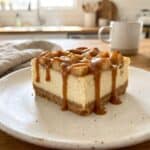 A caramel apple cheesecake bar on a plate drizzled with caramel sauce and topped with apples.