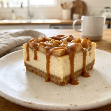A caramel apple cheesecake bar on a plate drizzled with caramel sauce and topped with apples.