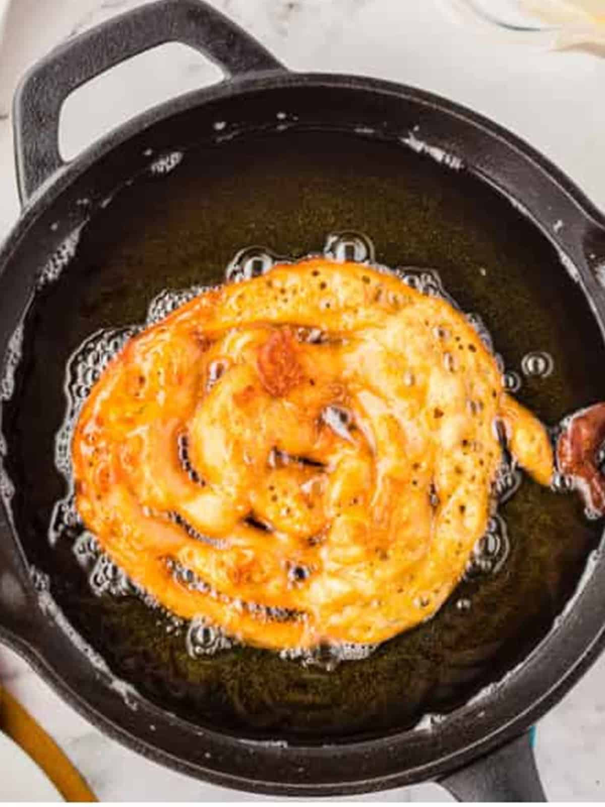 Funnel cake batter piped into hot oil and frying to golden brown in a cast iron skillet.