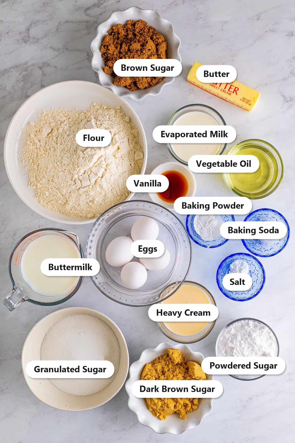 Labeled ingredients for homemade caramel cake including flour, granulated sugar, brown sugar, butter, eggs, buttermilk, oil, vanilla, evaporated milk, heavy cream, and powdered sugar.