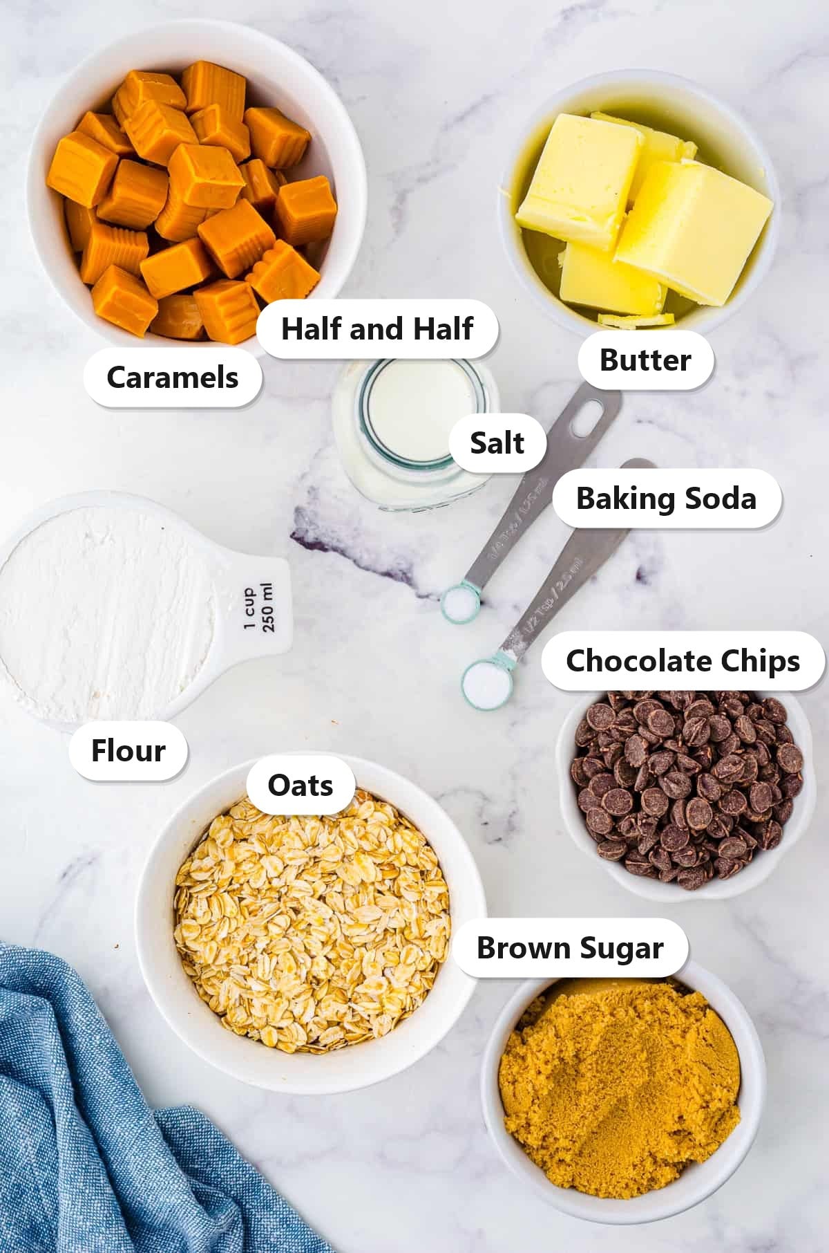 Carmelitas ingredients including caramels, butter, half and half, oats, flour, brown sugar, baking soda, salt, and chocolate chips labeled on a marble counter.