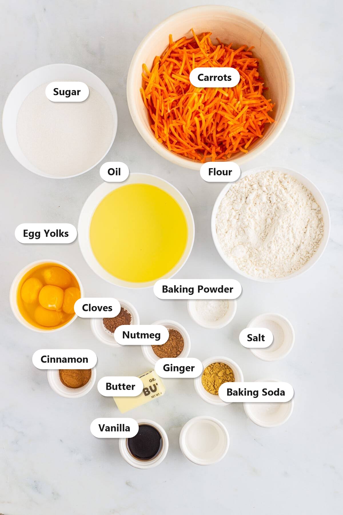 Labeled ingredients to make carrot bundt cake including grated carrots, cake flour, sugar, oil, butter, eggs, and warm spices like cinnamon, ginger, nutmeg, and cloves.