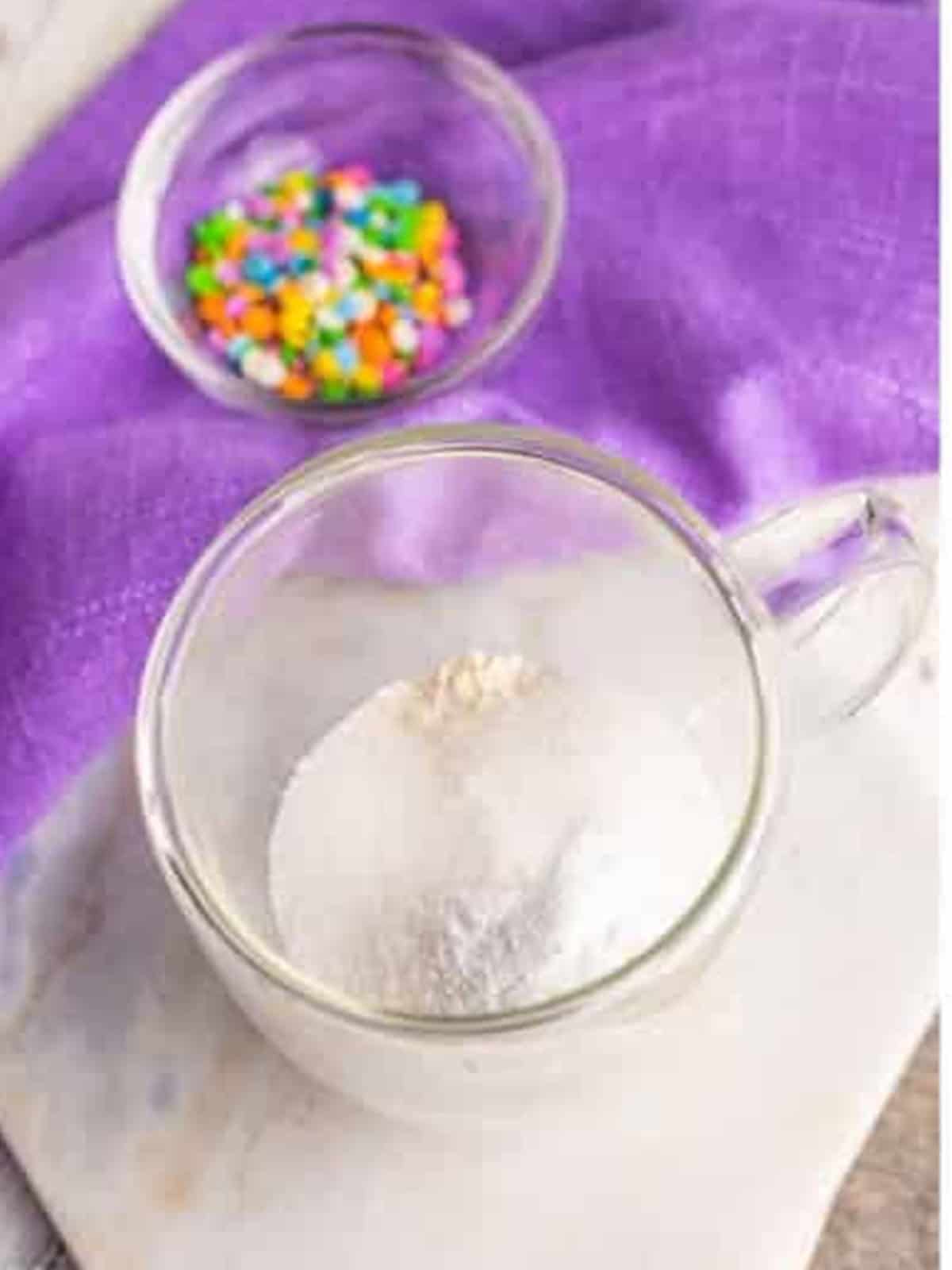 Adding flour, sugar, and baking powder to the bottom of a clear mug.