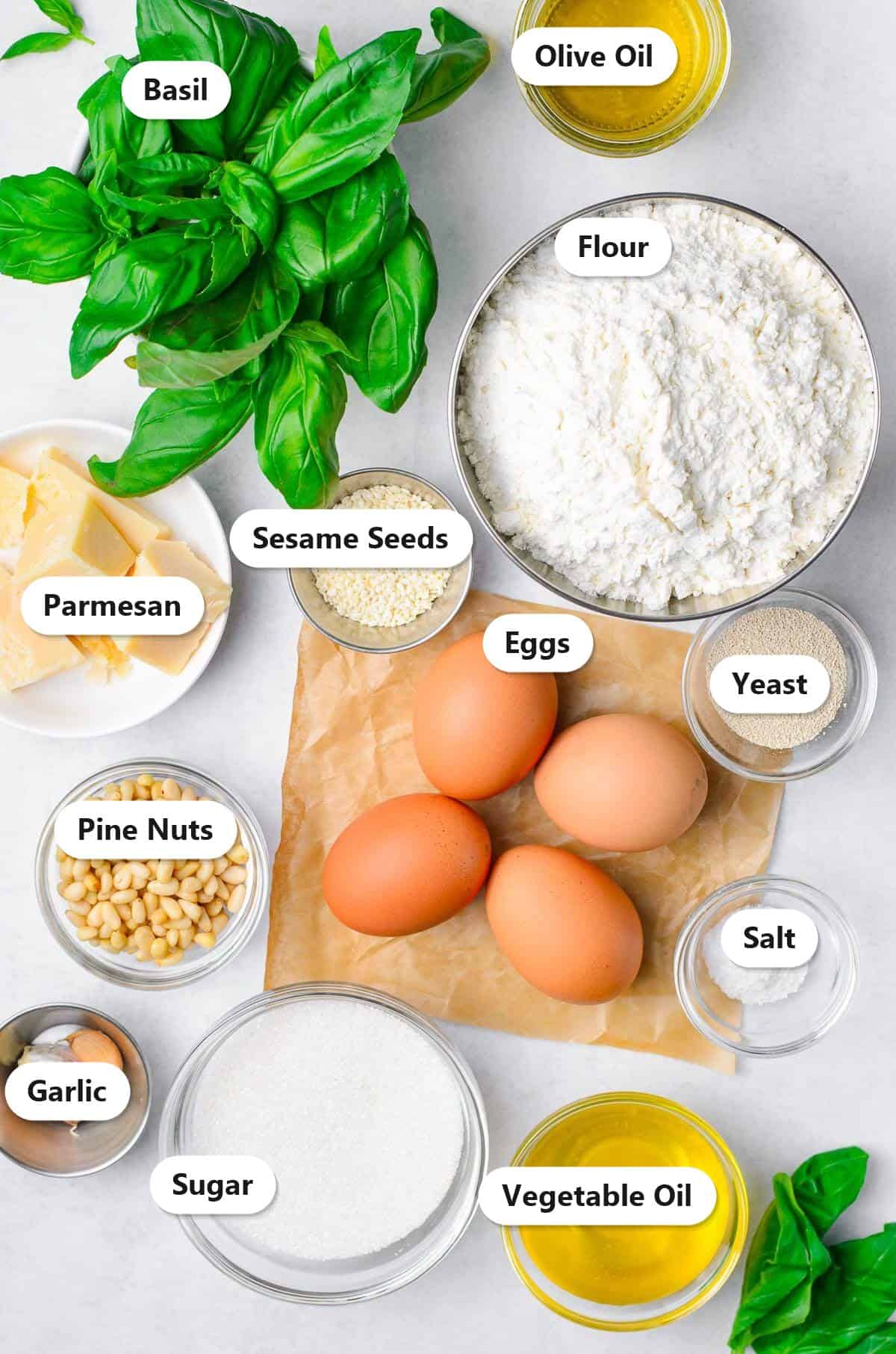 Labeled ingredients to make challah bread including flour, eggs, yeast, sugar, vegetable oil, salt, basil, parmesan, pine nuts, garlic, olive oil, and sesame seeds.
