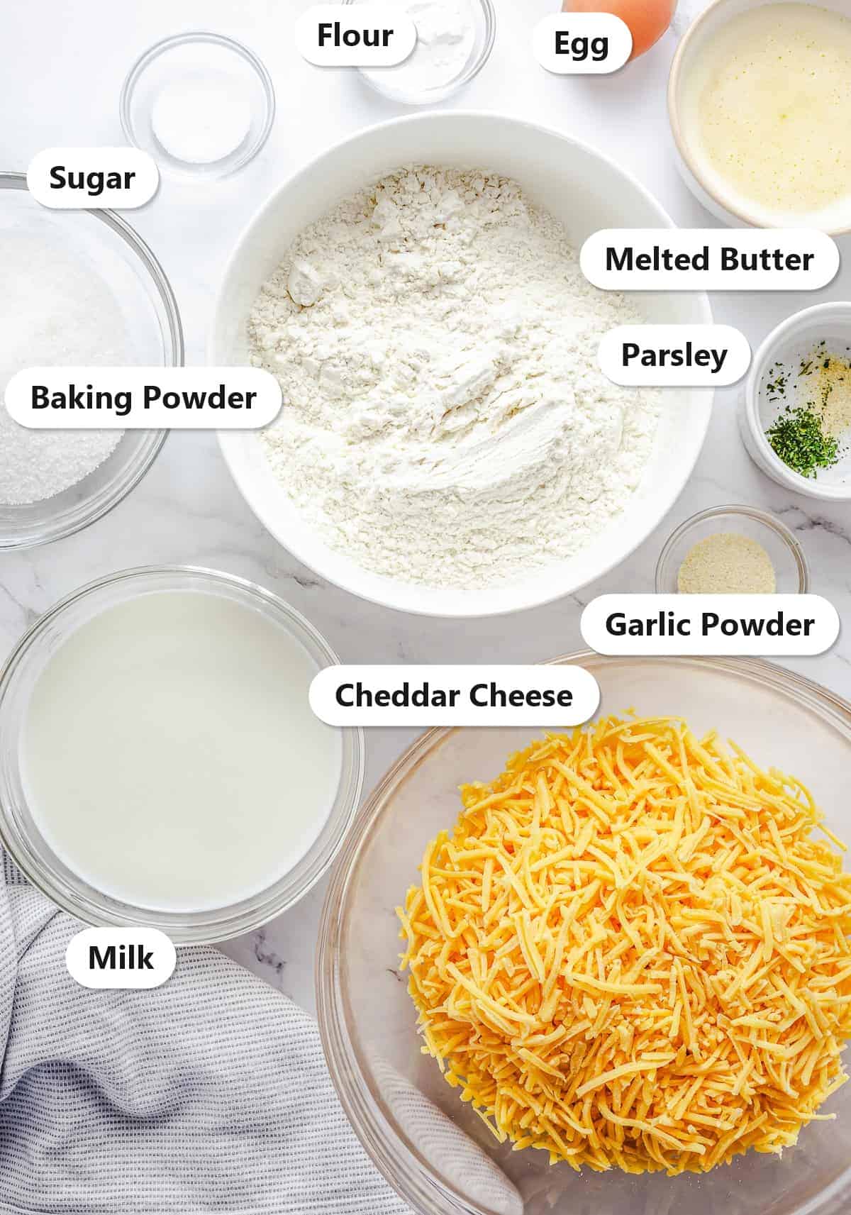 Labeled ingredients for cheese muffins including flour, cheddar cheese, milk, egg, melted butter, sugar, baking powder, parsley, and garlic powder.