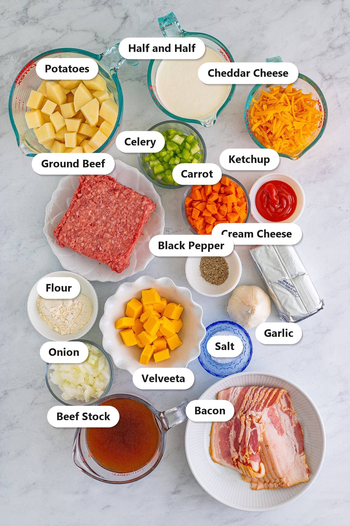 Labeled ingredients for cheeseburger soup including ground beef, bacon, potatoes, onion, carrot, celery, garlic, flour, beef stock, half and half, ketchup, cream cheese, Velveeta, cheddar cheese, salt, and black pepper.