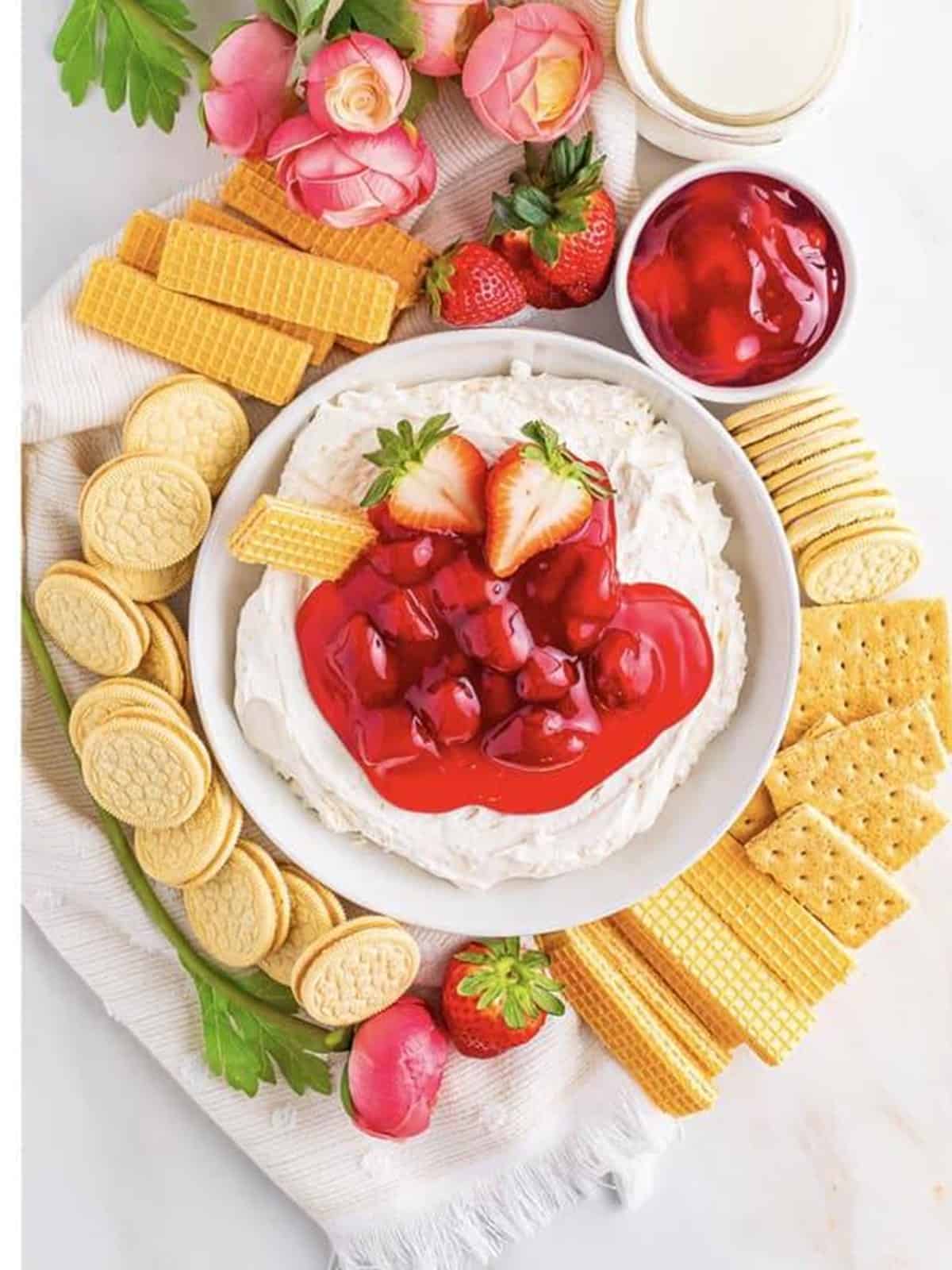 Finished cheesecake dip on a dessert board topped with strawberry pie filling and surrounded by cookies.