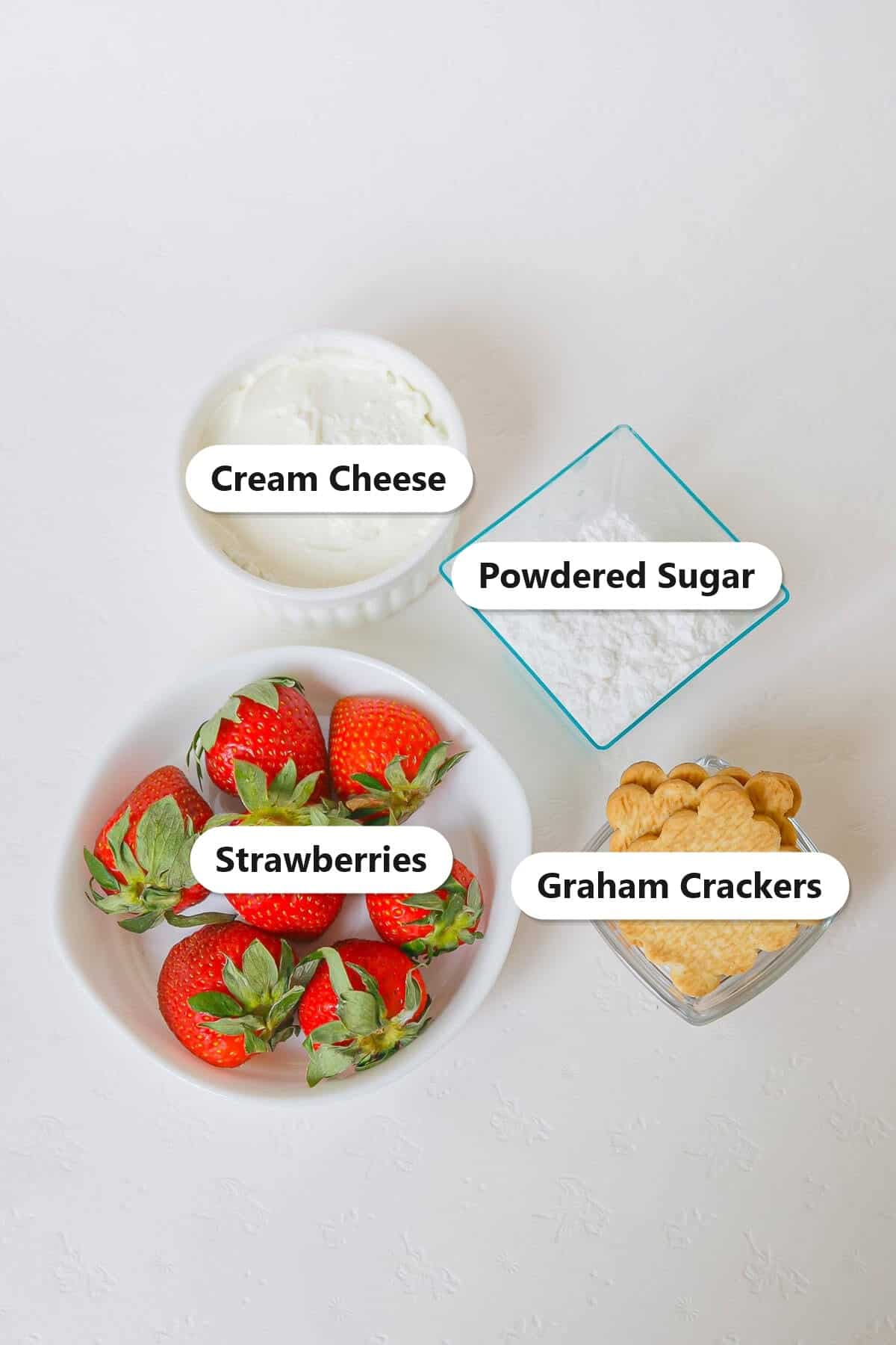 Labeled ingredients for cheesecake stuffed strawberries including fresh strawberries, cream cheese, powdered sugar, and graham crackers.