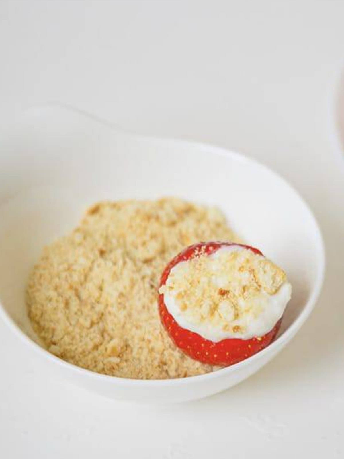 A cheesecake stuffed strawberry being dipped into graham cracker crumbs.