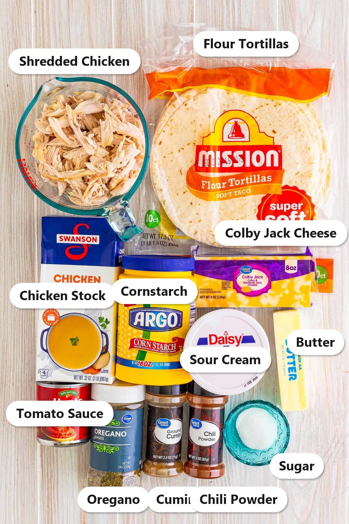 Labeled ingredients to make chicken enchiladas including rotisserie chicken, flour tortillas, chicken stock, cornstarch, colby jack cheese, sour cream, butter, tomato sauce, and spices.