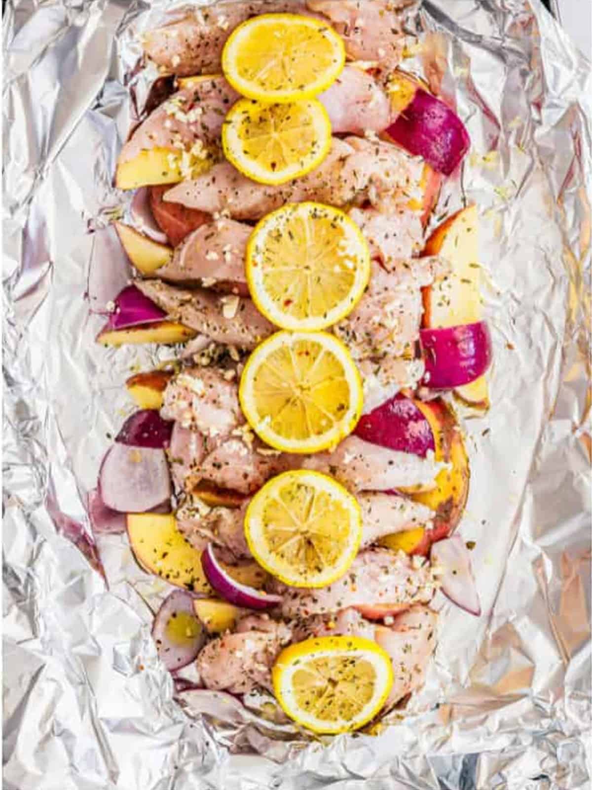 Seasoned chicken, potatoes, and onion arranged on foil and topped with thin lemon slices.