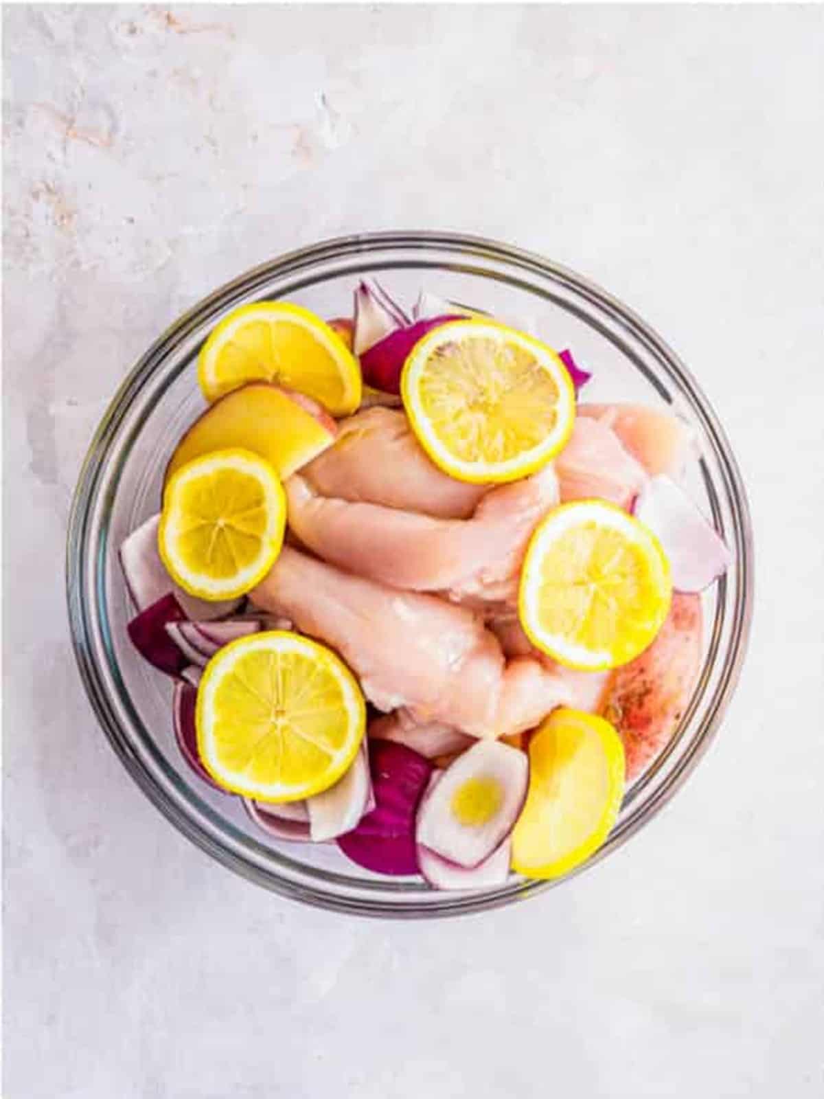 Chicken tenderloins, red potatoes, red onion, and lemon slices tossed with olive oil in a glass bowl.