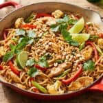 Chicken pad thai in a red skillet topped with peanuts, cilantro, mint, and lime wedges.