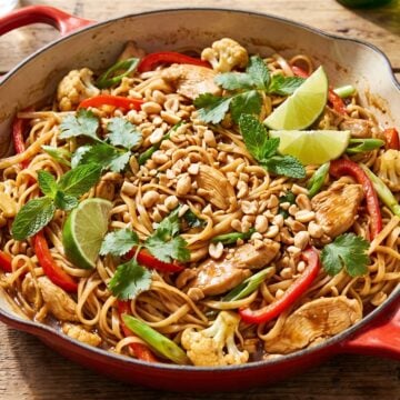 Chicken pad thai in a red skillet topped with peanuts, cilantro, mint, and lime wedges.