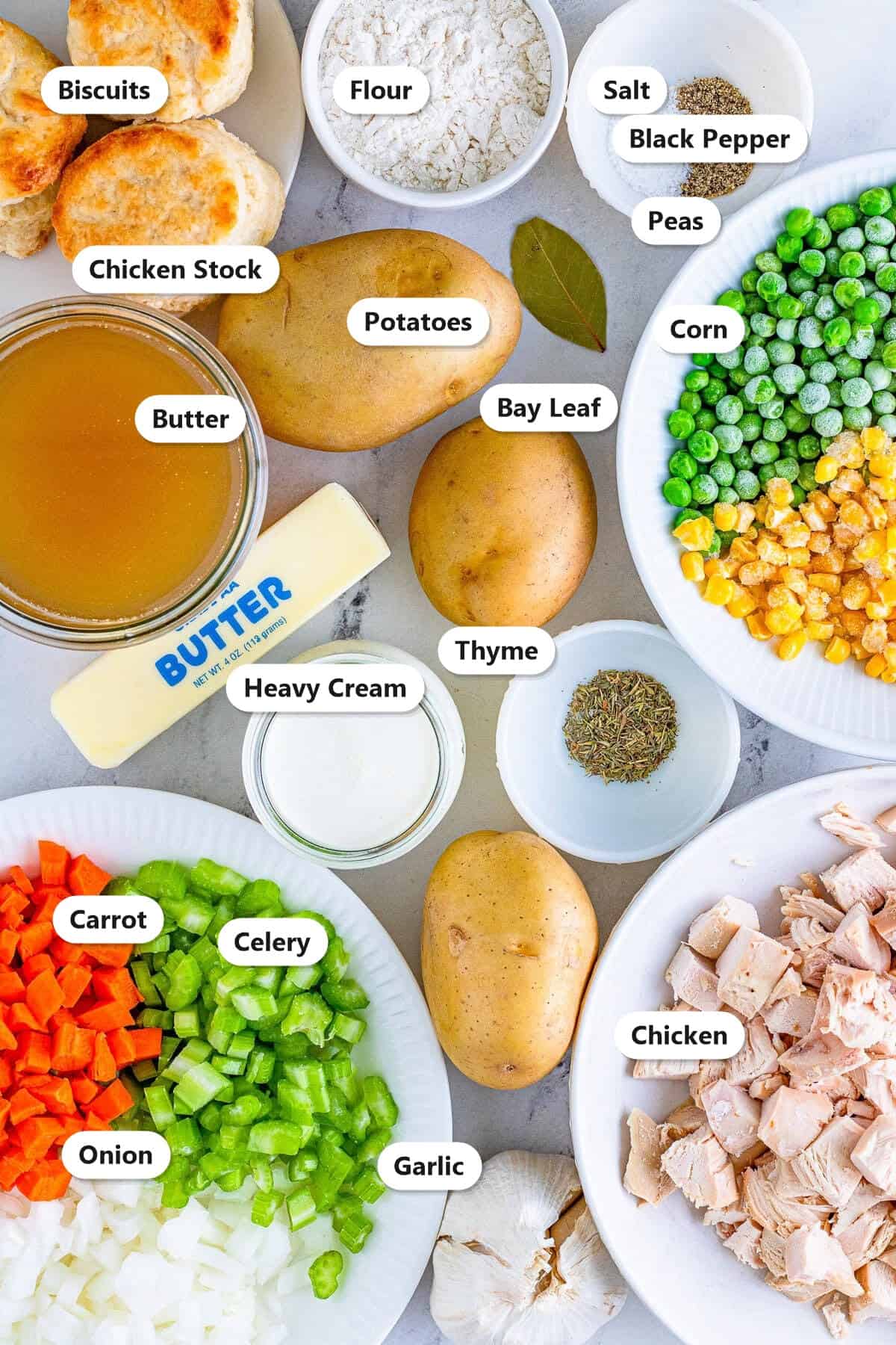 Chicken pot pie soup ingredients labeled on a marble surface including butter, chicken stock, potatoes, carrot, celery, onion, garlic, peas, corn, cooked chicken, heavy cream, flour, thyme, bay leaf, salt, and black pepper.