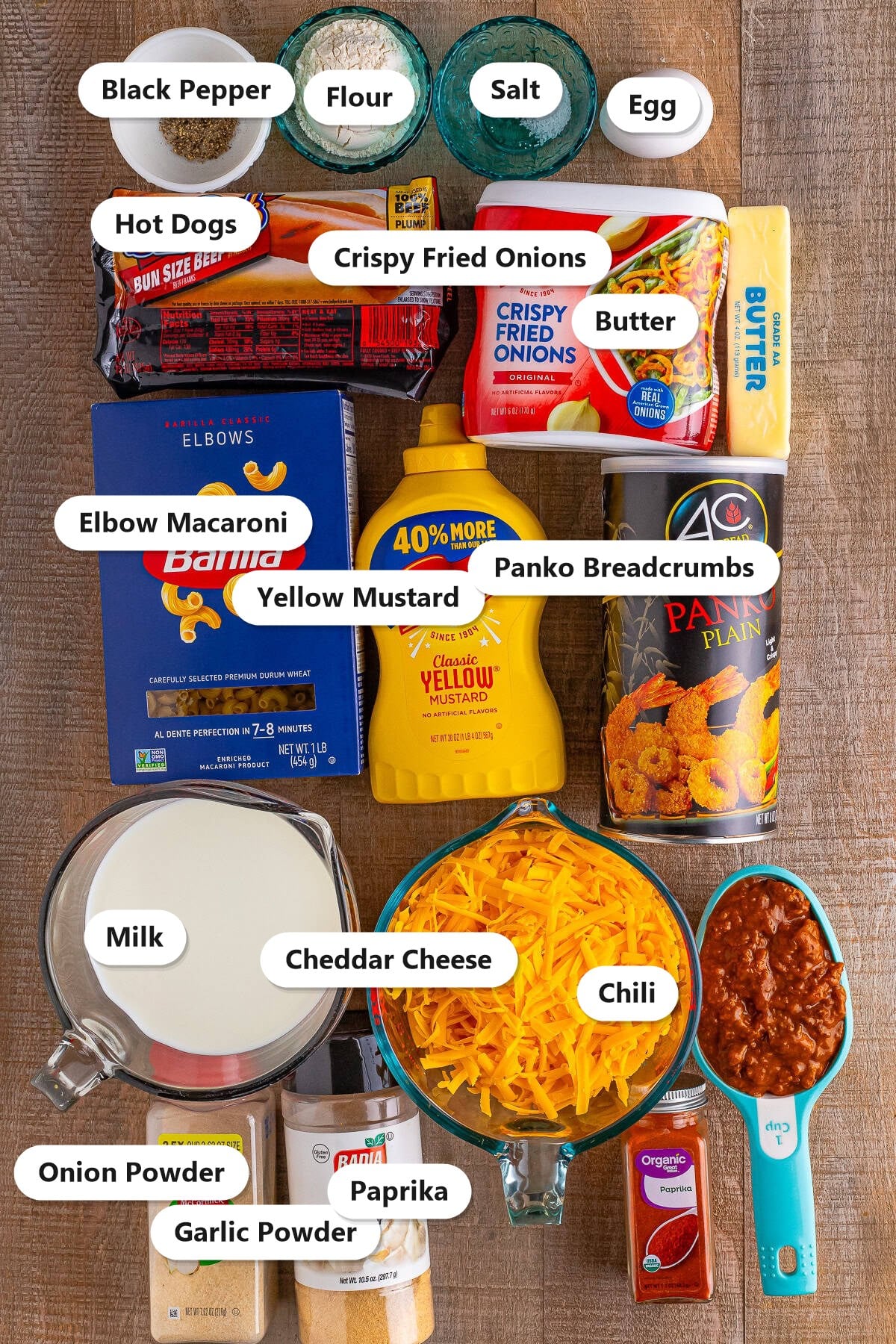Ingredients to make chili cheese dog mac and cheese labeled on a wood surface including hot dogs, elbow macaroni, cheddar, chili, crispy fried onions, panko, milk, butter, mustard, and seasonings.