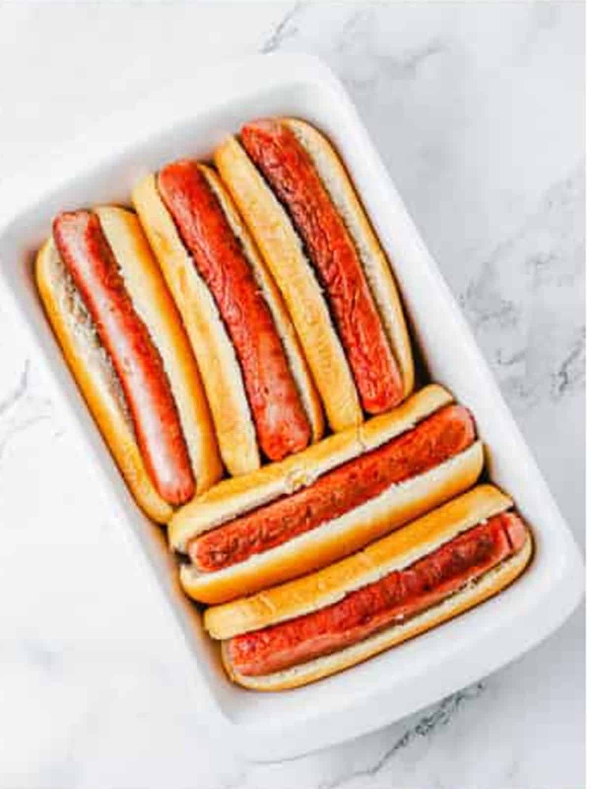 Hot dogs nestled into buns in a white baking dish.