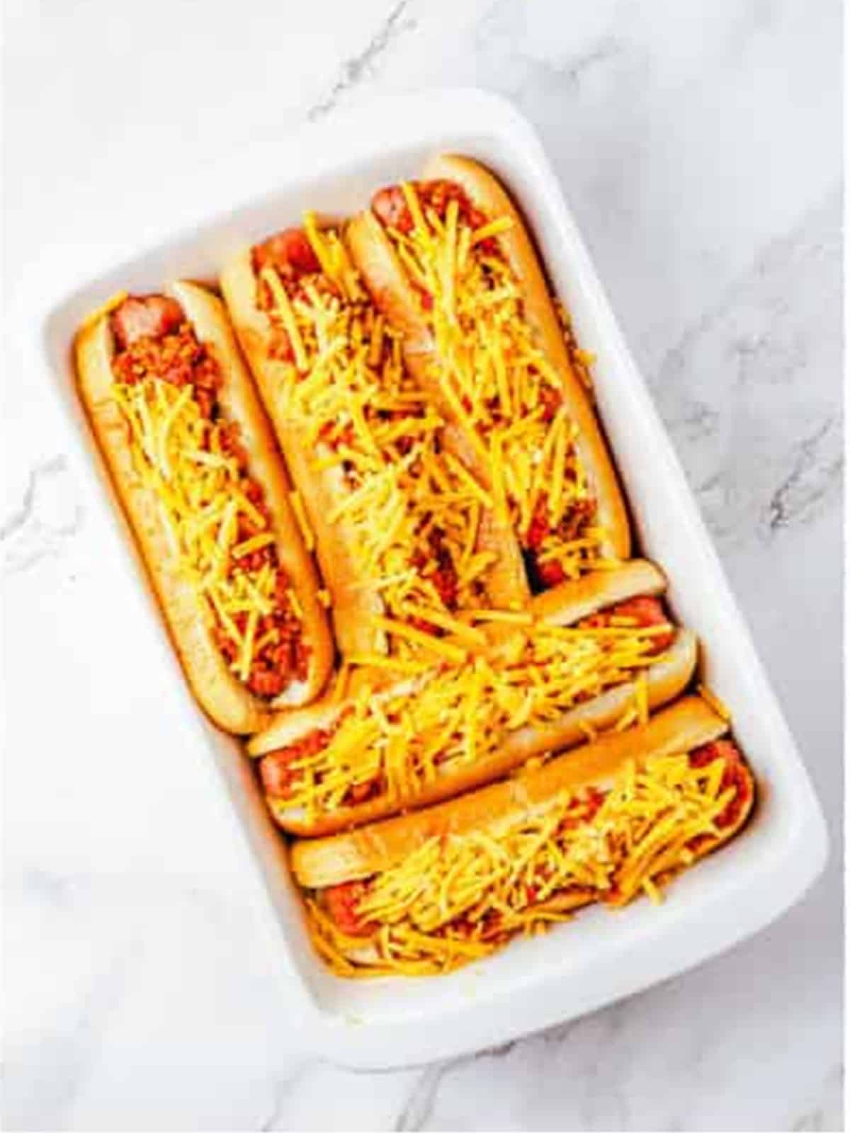 Hot dogs topped with chili sauce and shredded cheddar in a baking dish.