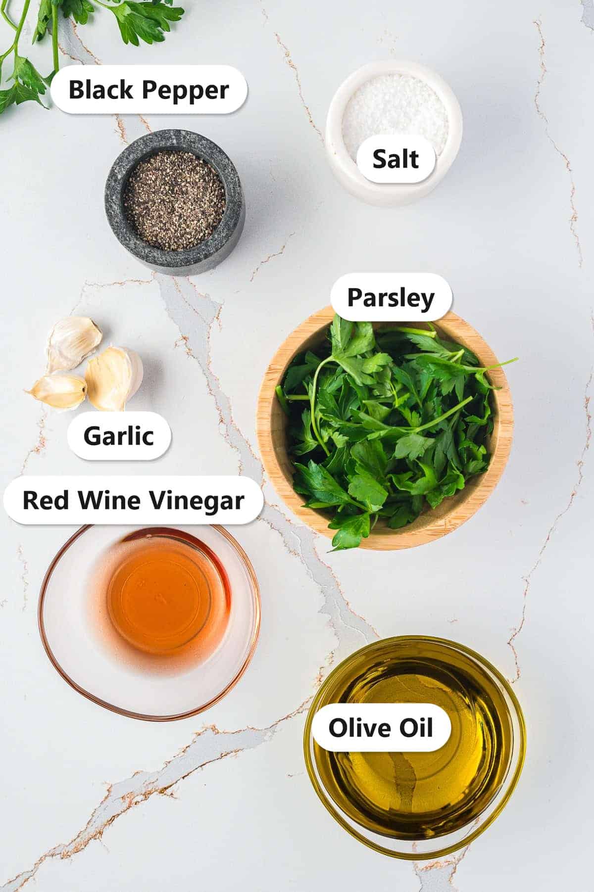 Labeled ingredients for chimichurri sauce including parsley, red wine vinegar, salt, black pepper, garlic, and olive oil.