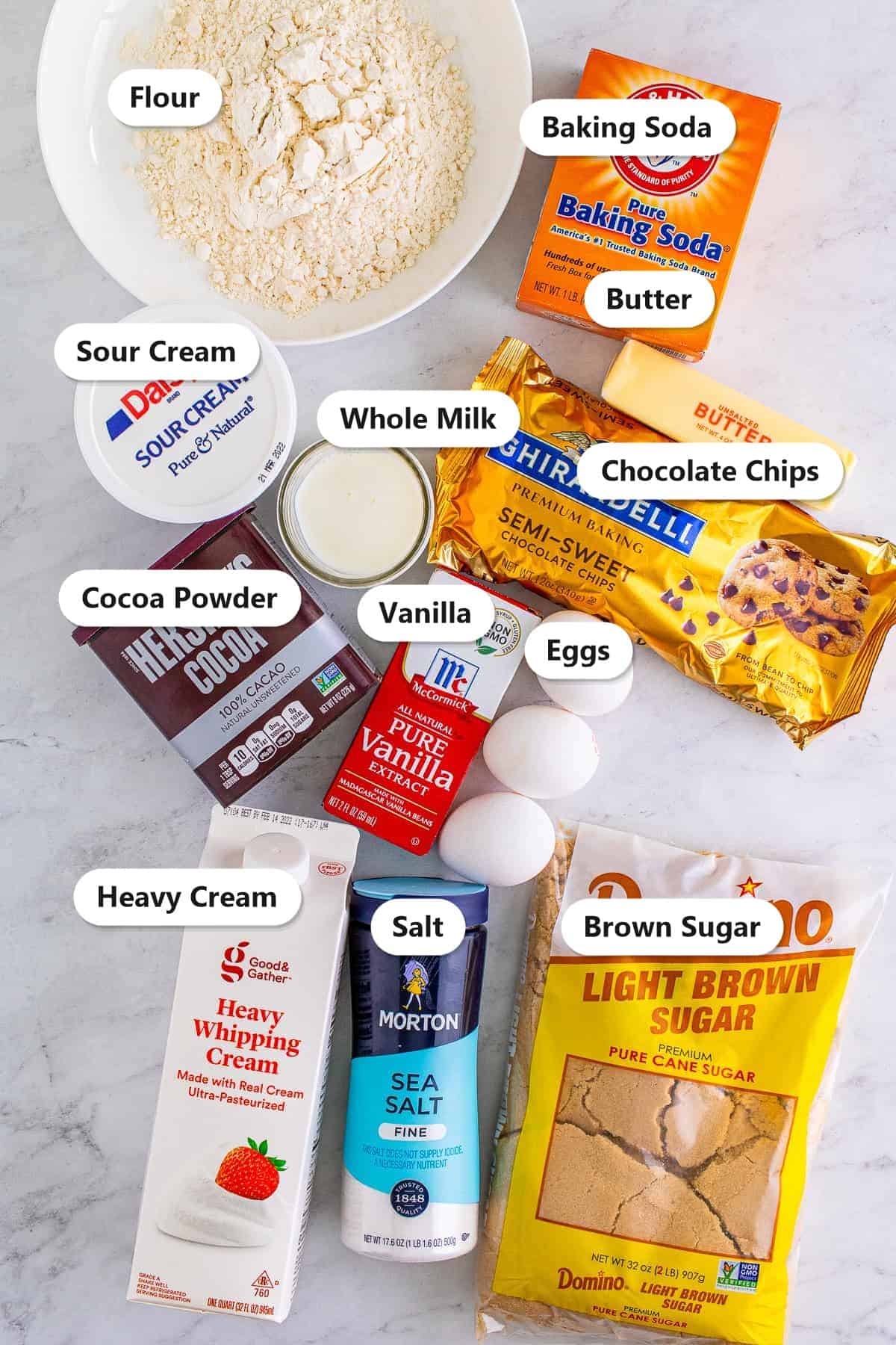 Labeled ingredients to make chocolate bundt cake including flour, cocoa powder, chocolate chips, sour cream, butter, brown sugar, eggs, and heavy cream.