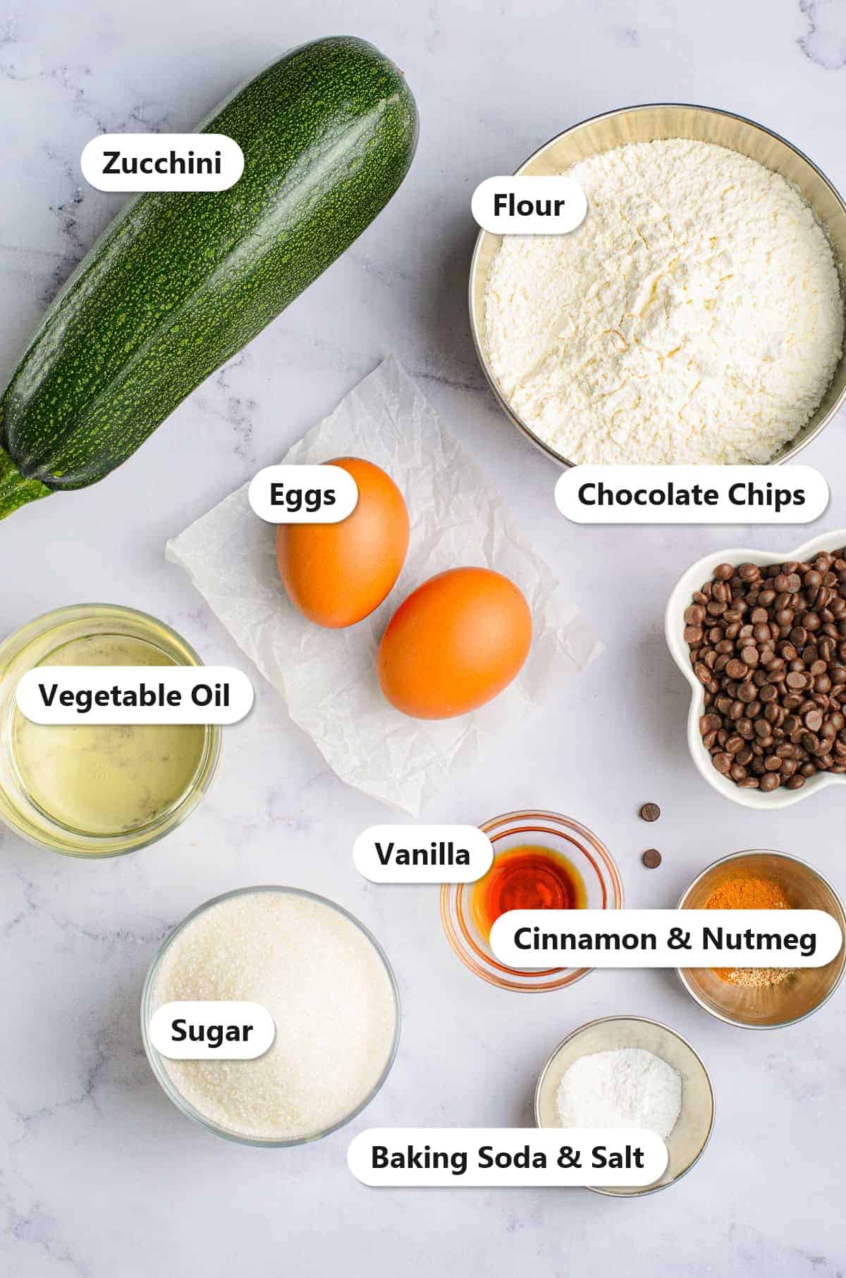 Labeled ingredients for chocolate chip zucchini bread including zucchini, flour, eggs, oil, sugar, chocolate chips, and spices.