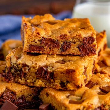 A stack of chocolate chunk blondies showing gooey melted chocolate, next to a jar of milk.