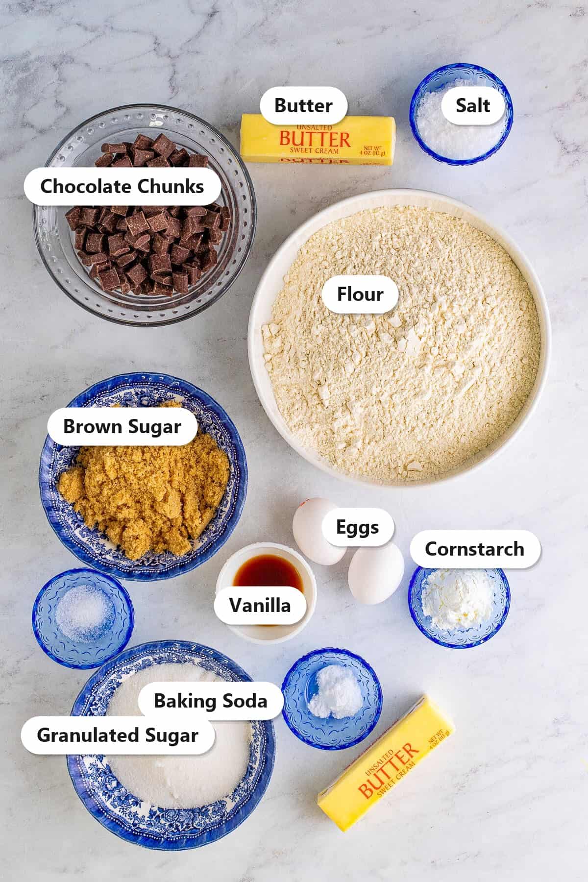 Labeled ingredients to make chocolate chunk cookies including chocolate chunks, flour, brown sugar, butter, eggs, cornstarch, and vanilla.