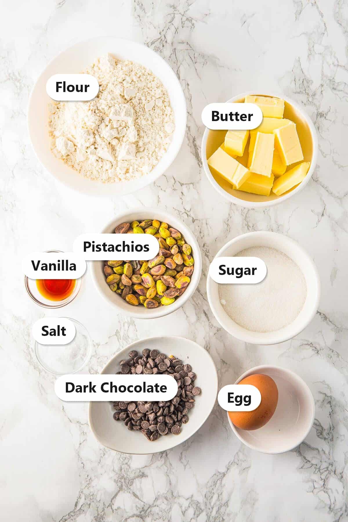 Labeled ingredients for Chocolate Dipped Pistachio Cookies including flour, butter, pistachios, sugar, egg, vanilla, and dark chocolate.