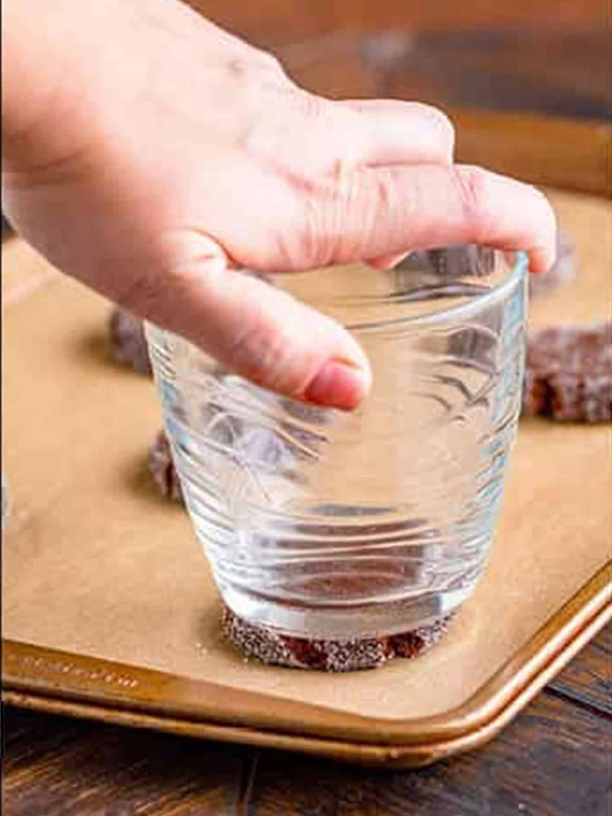Flattening a sugar-coated chocolate cookie dough ball with the bottom of a glass.