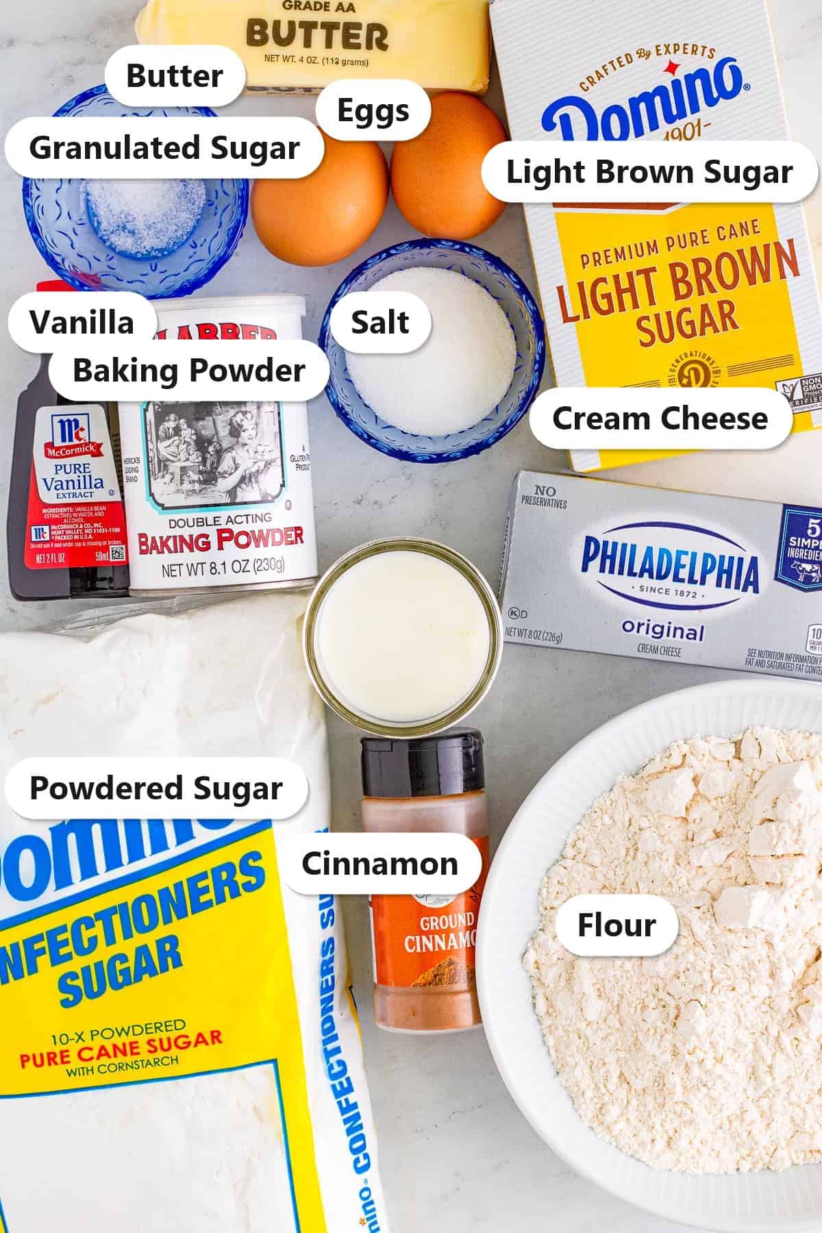 Labeled ingredients to make cinnamon roll blondies including flour, brown sugar, butter, eggs, vanilla, cinnamon, cream cheese, and powdered sugar.