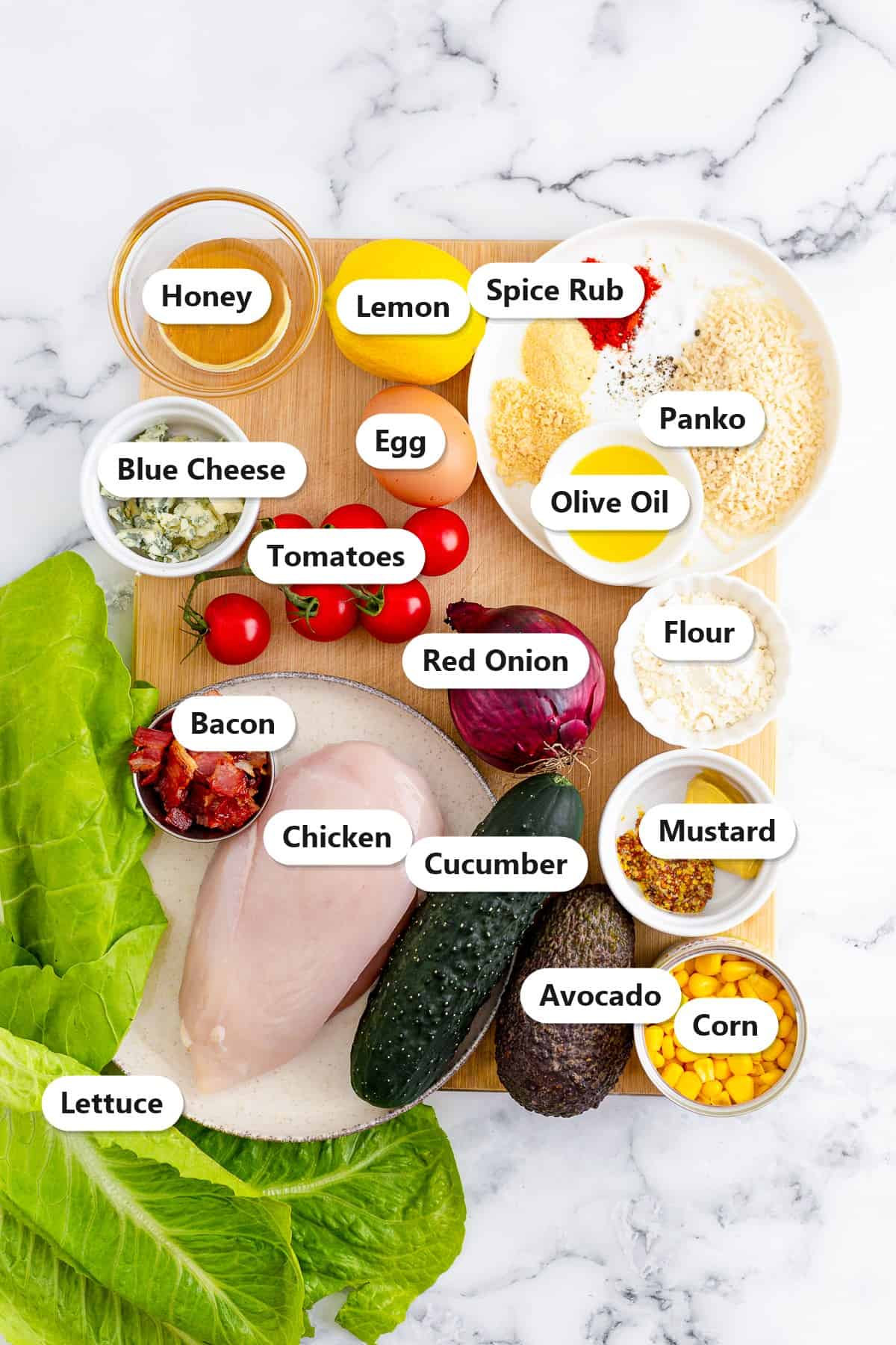Cobb Salad ingredients including chicken, lettuce, bacon, blue cheese, egg, tomatoes, avocado, corn, cucumber, red onion, and honey mustard labeled on a cutting board.