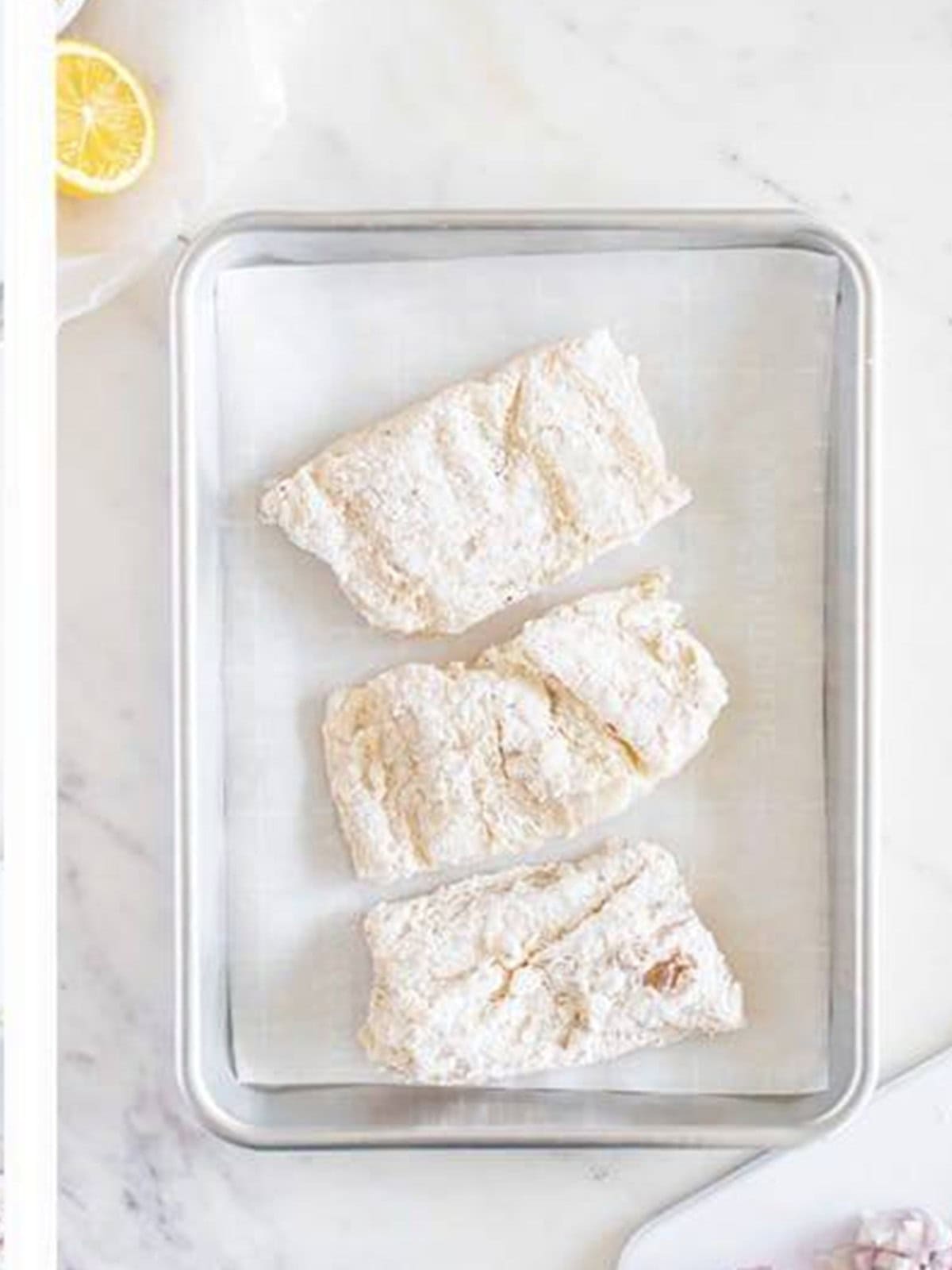 Floured cod fillets resting on a sheet tray before pan-frying.