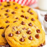 A stack of copycat Mrs Fields chocolate chip cookies on a white plate with a red checkered napkin and a glass of milk.