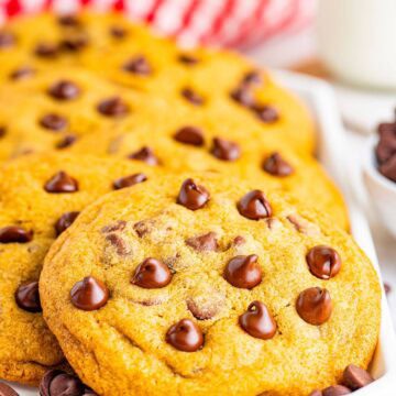 A stack of copycat Mrs Fields chocolate chip cookies on a white plate with a red checkered napkin and a glass of milk.