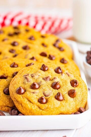 A stack of copycat Mrs Fields chocolate chip cookies on a white plate with a red checkered napkin and a glass of milk.