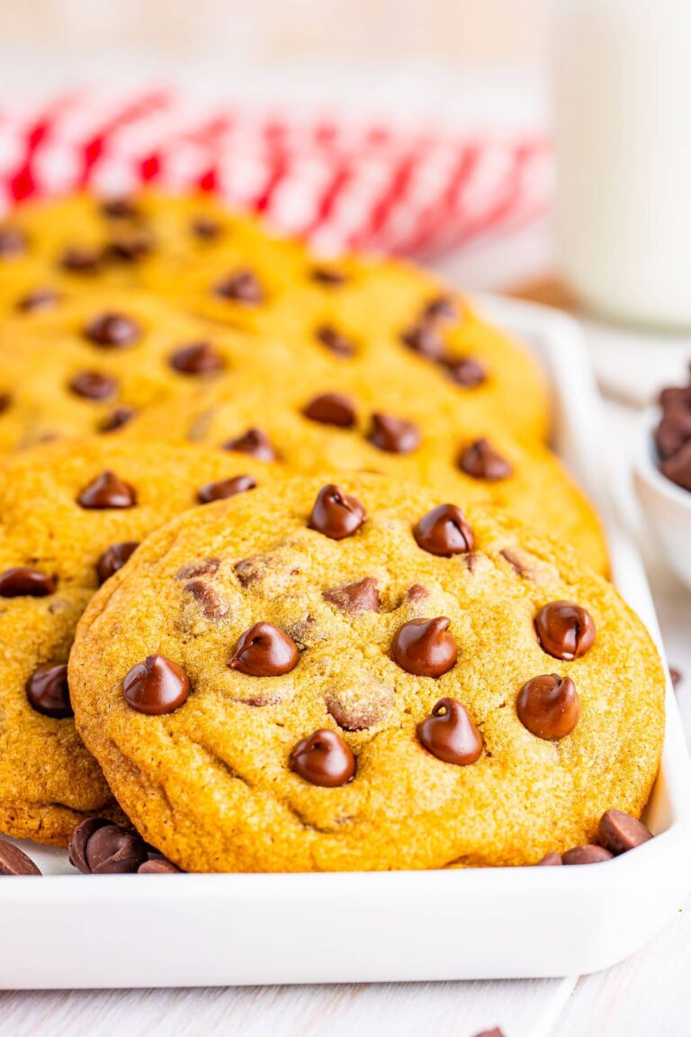 A stack of copycat Mrs Fields chocolate chip cookies on a white plate with a red checkered napkin and a glass of milk.