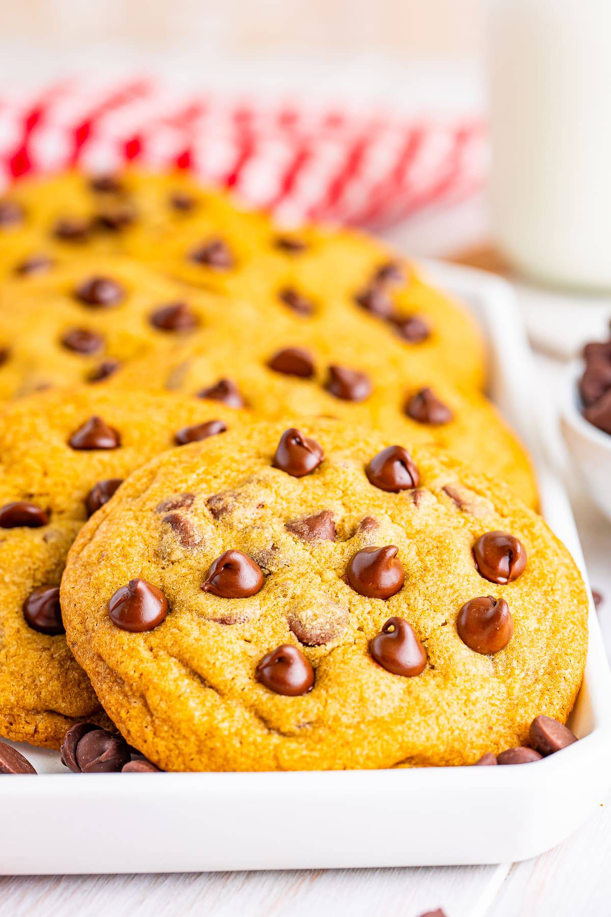 A stack of copycat Mrs Fields chocolate chip cookies on a white plate with a red checkered napkin and a glass of milk.