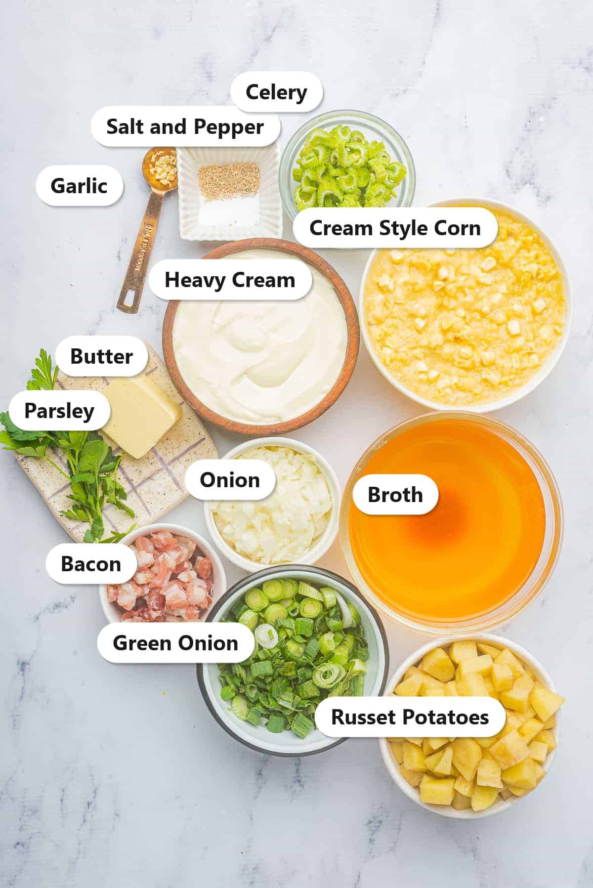 Ingredients to make corn chowder labeled on a marble surface: bacon, onion, garlic, celery, green onion, russet potatoes, cream style corn, broth, heavy cream, butter, and salt and pepper.