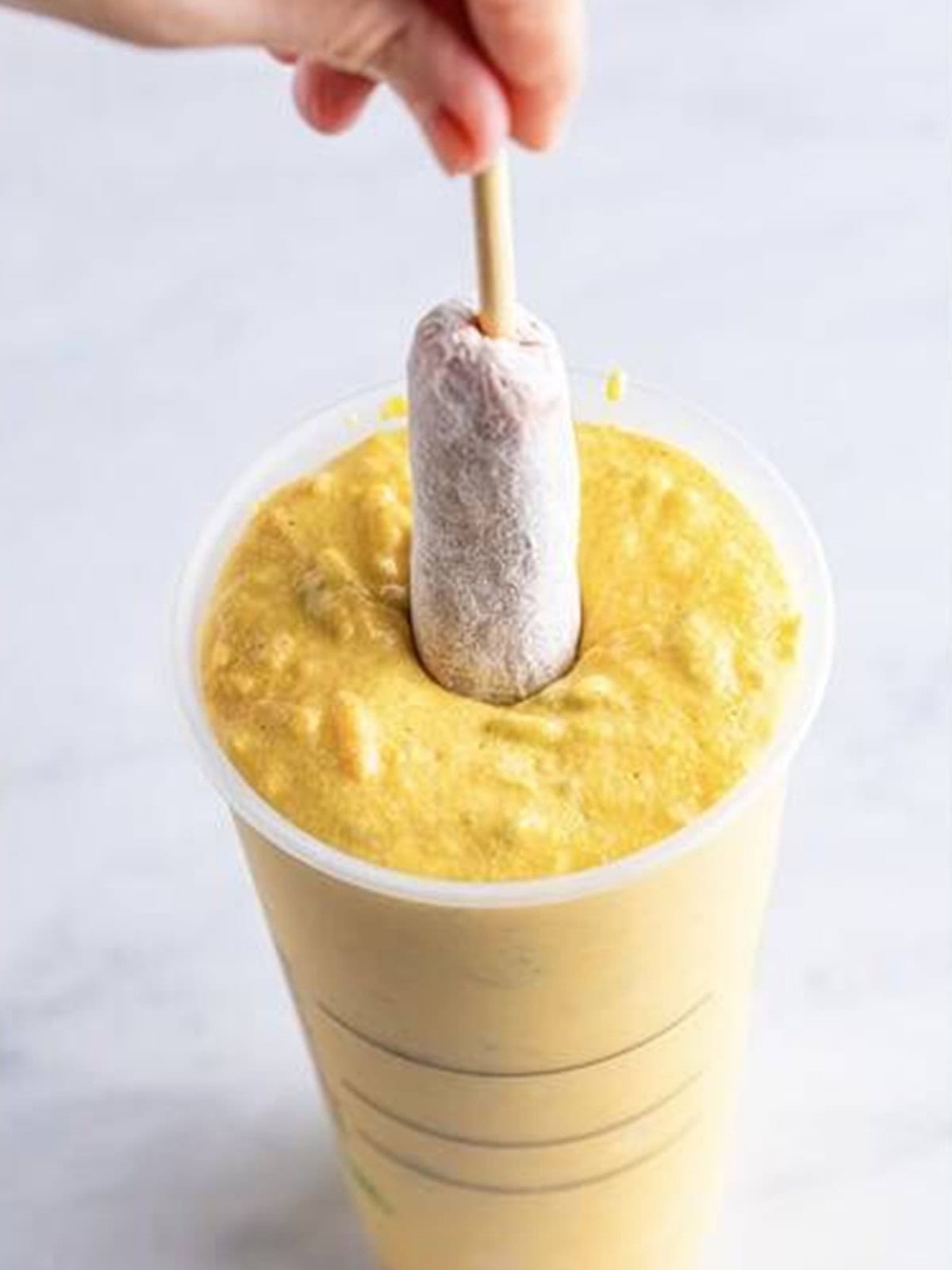 A skewered hot dog dipped into corn dog batter in a tall cup.