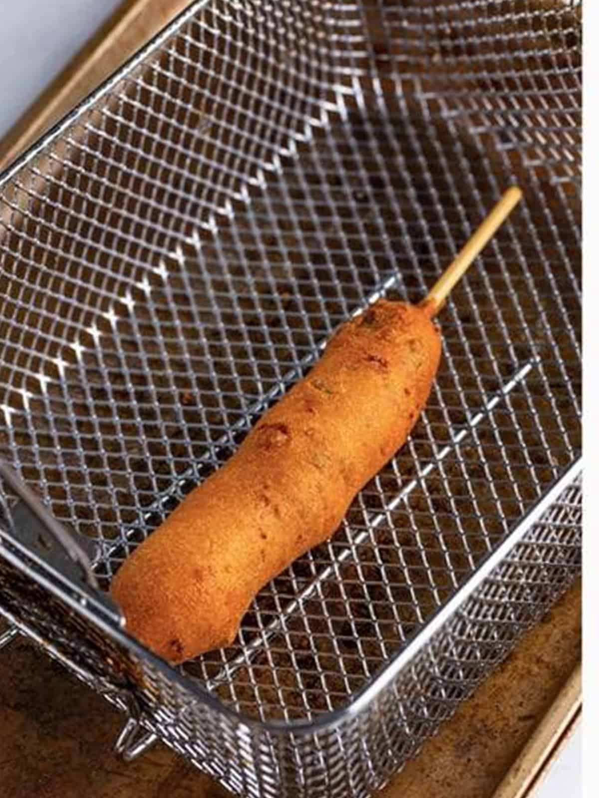 A battered corn dog frying golden in a fryer basket.