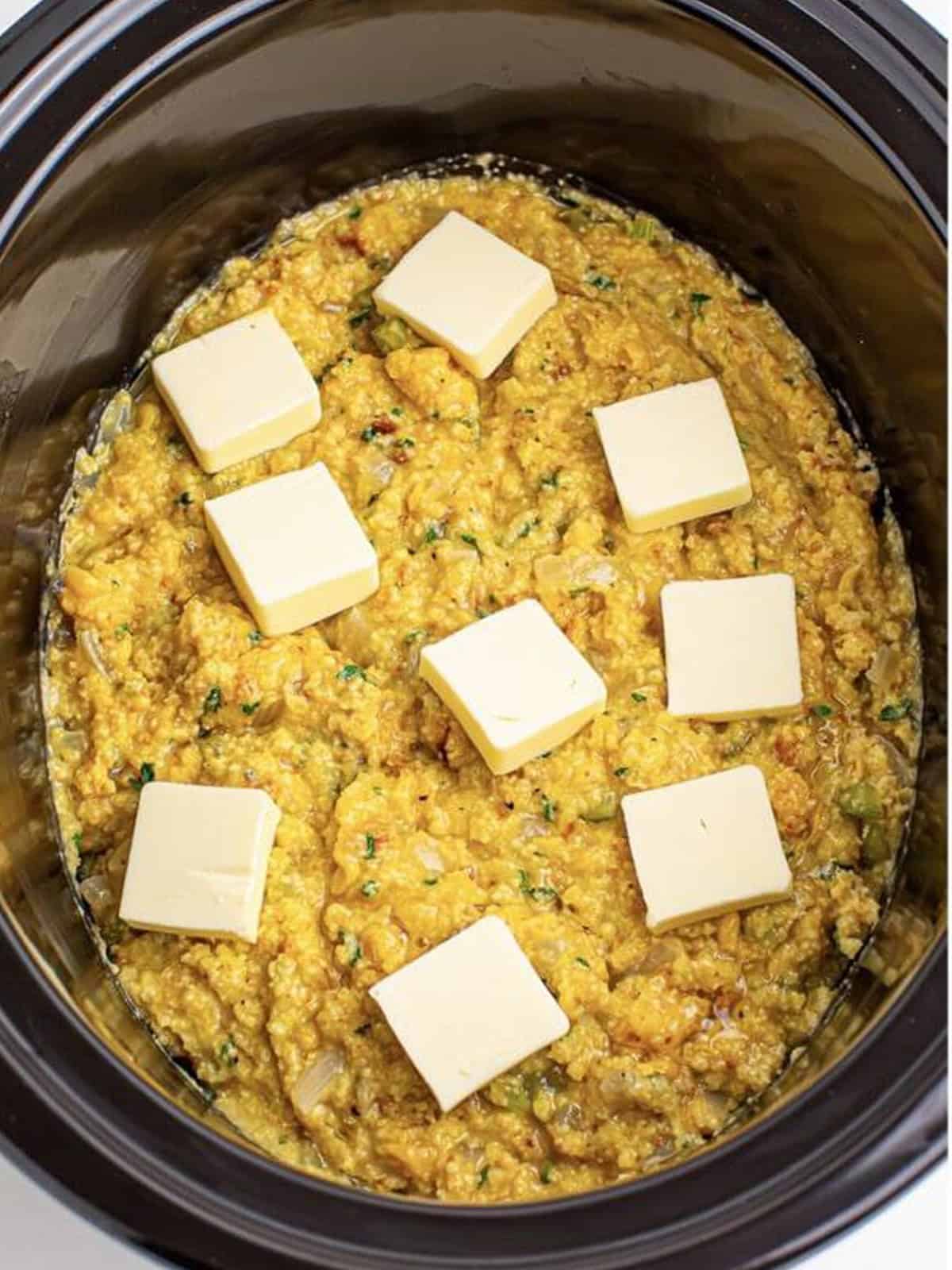 Pats of butter dotted across the top of the cornbread dressing in the slow cooker.