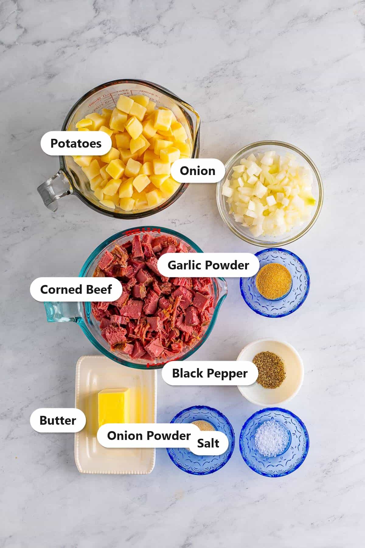 Corned beef hash ingredients including cooked potatoes, corned beef, onion, butter, garlic powder, onion powder, salt, and pepper labeled on a marble surface.