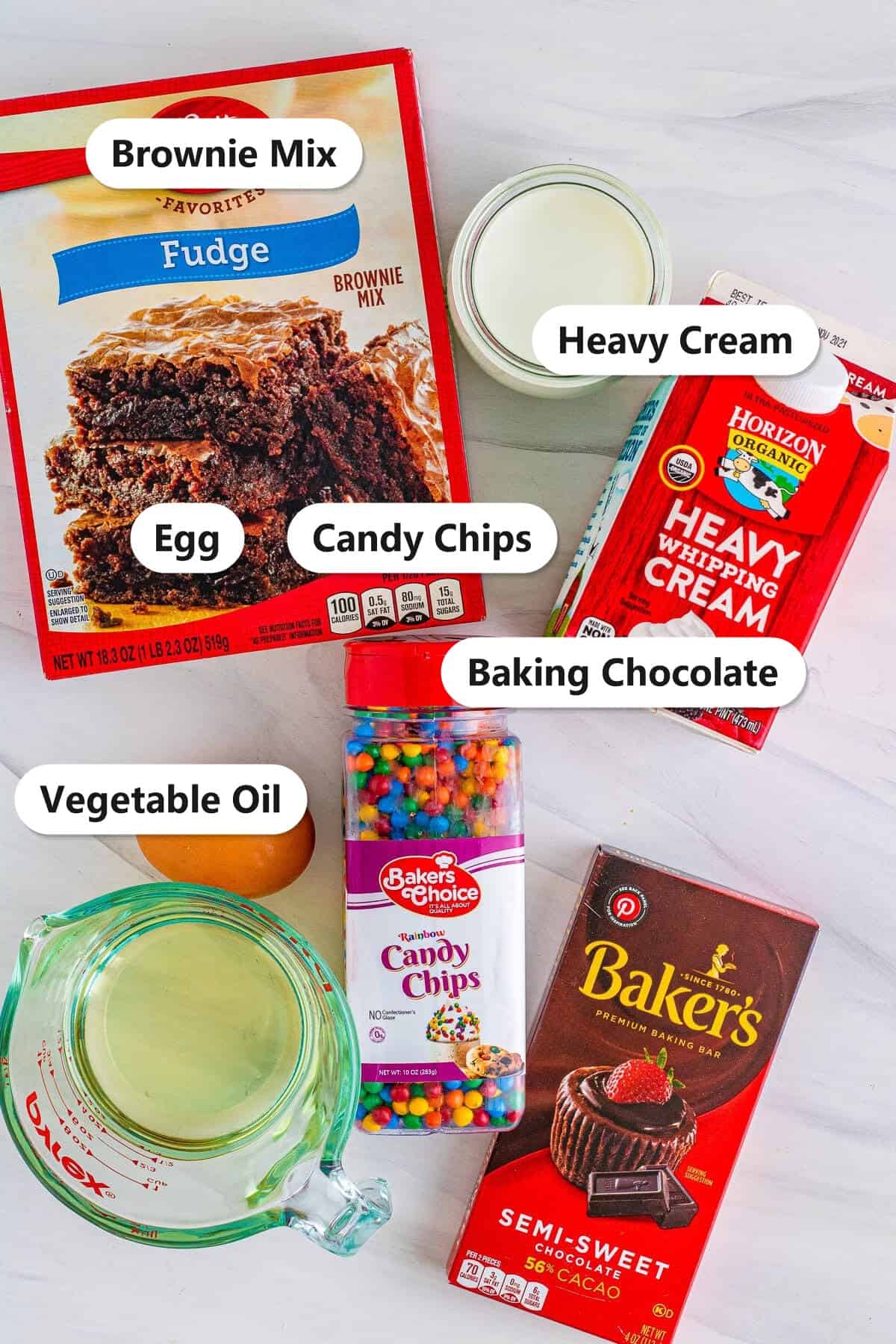 Labeled ingredients to make cosmic brownie cookies including brownie mix, heavy cream, egg, vegetable oil, candy chips, and baking chocolate.