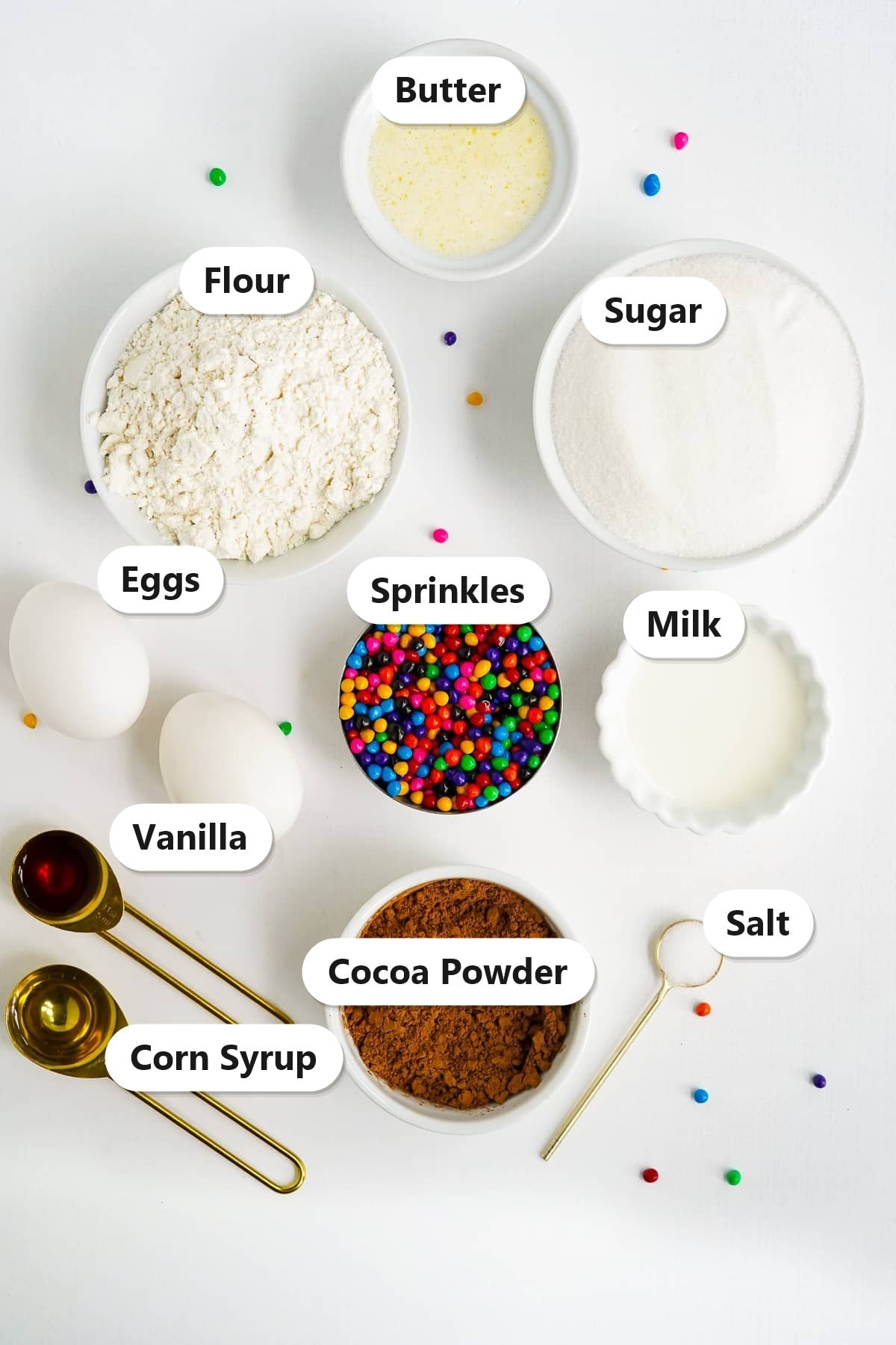 Labeled ingredients to make cosmic brownies including flour, granulated sugar, cocoa powder, melted butter, eggs, milk, vanilla, corn syrup, salt, and rainbow chip sprinkles.