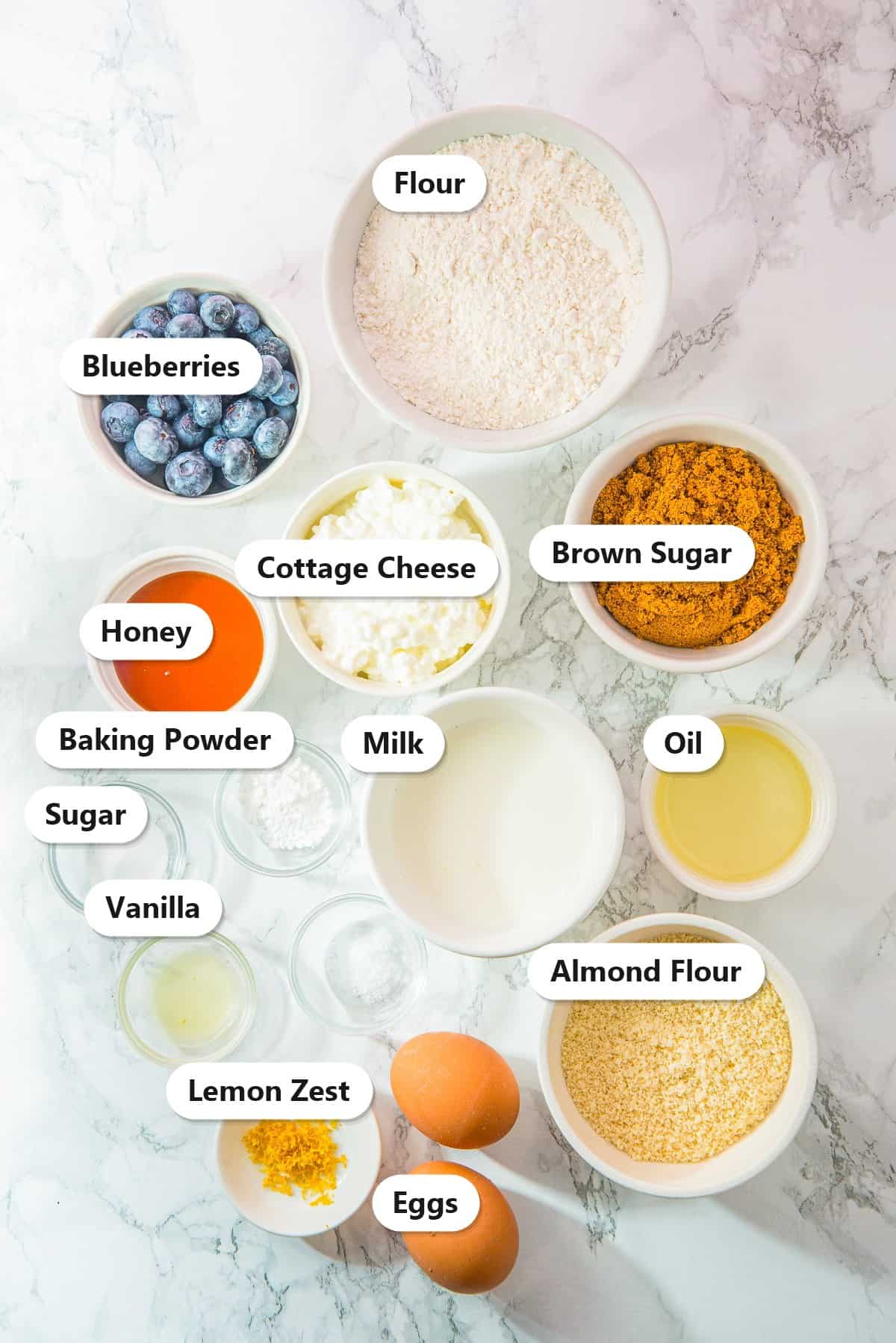 Labeled ingredients for Cottage Cheese Blueberry Muffins, including flour, blueberries, cottage cheese, brown sugar, honey, milk, oil, baking powder, sugar, vanilla, almond flour, lemon zest, and eggs.