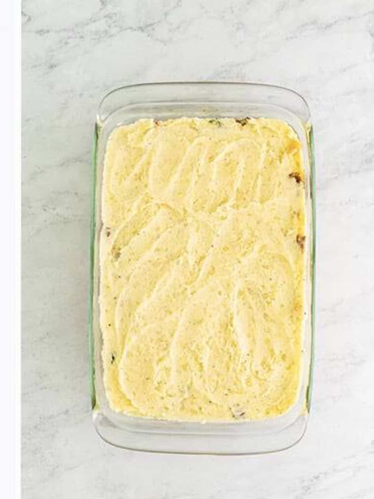 Mashed potatoes smoothed over the beef filling in a cottage pie baking dish.