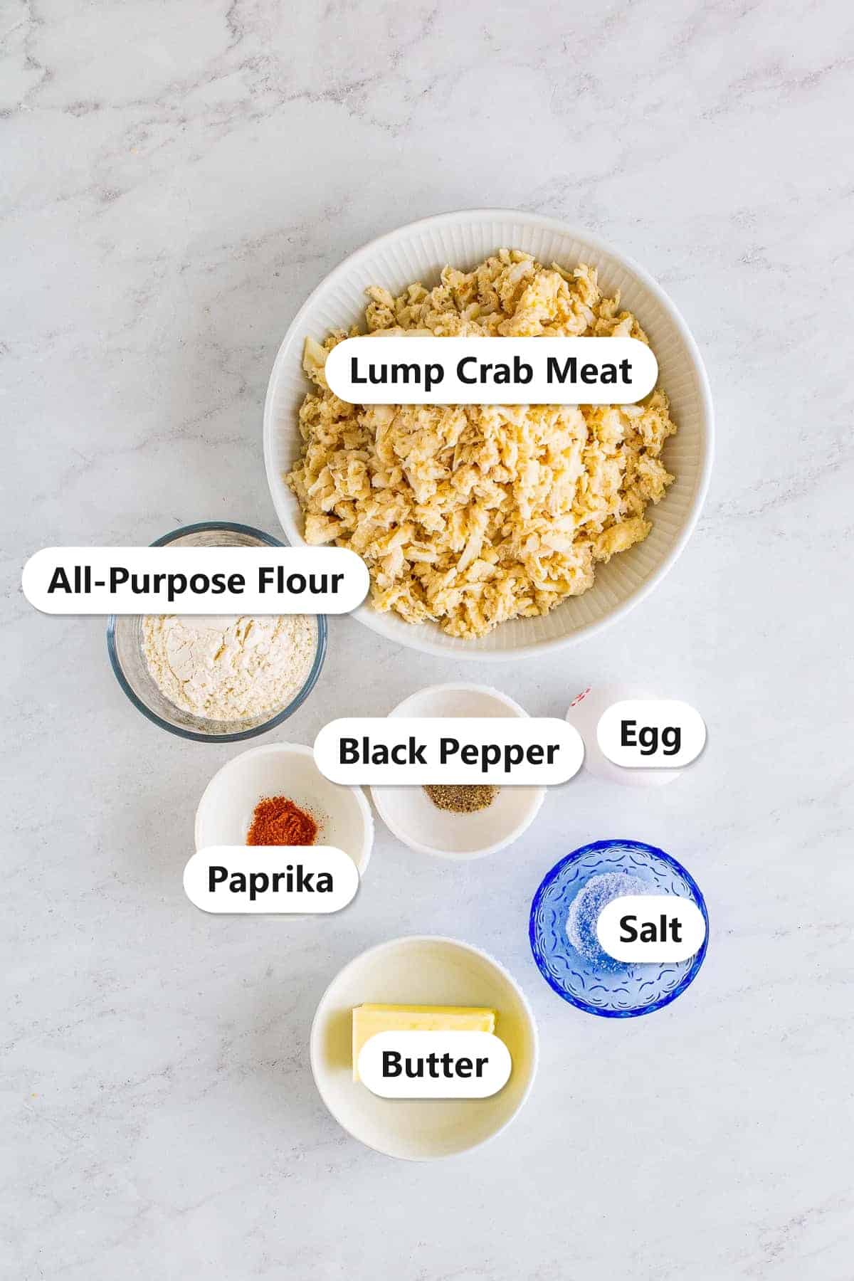 Labeled ingredients to make crab cakes including lump crab meat, flour, egg, paprika, salt, and butter.