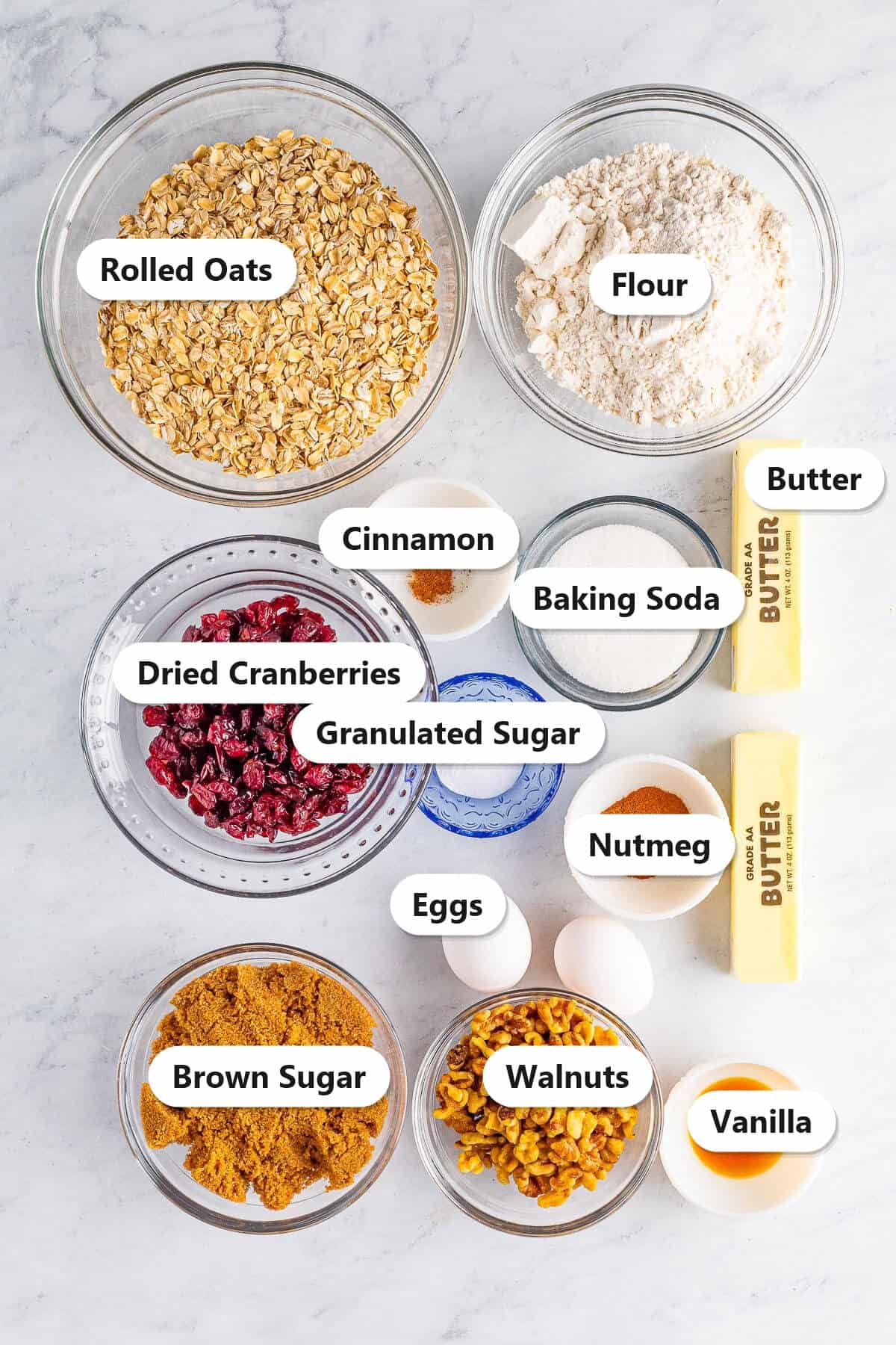 Cranberry oatmeal cookies ingredients including rolled oats, flour, dried cranberries, walnuts, butter, brown sugar, and eggs labeled on a marble surface.