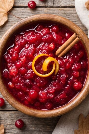 An overhead wooden bowl of homemade cranberry sauce with a cinnamon stick and orange peel, fall styling.